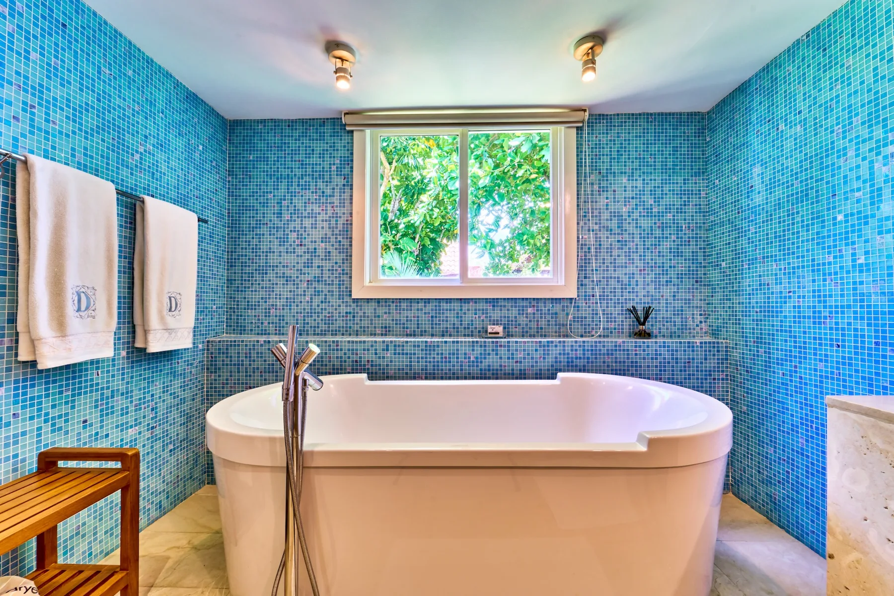 A bathroom with a freestanding bathtub, blue tiled walls, and a window overlooking greenery.