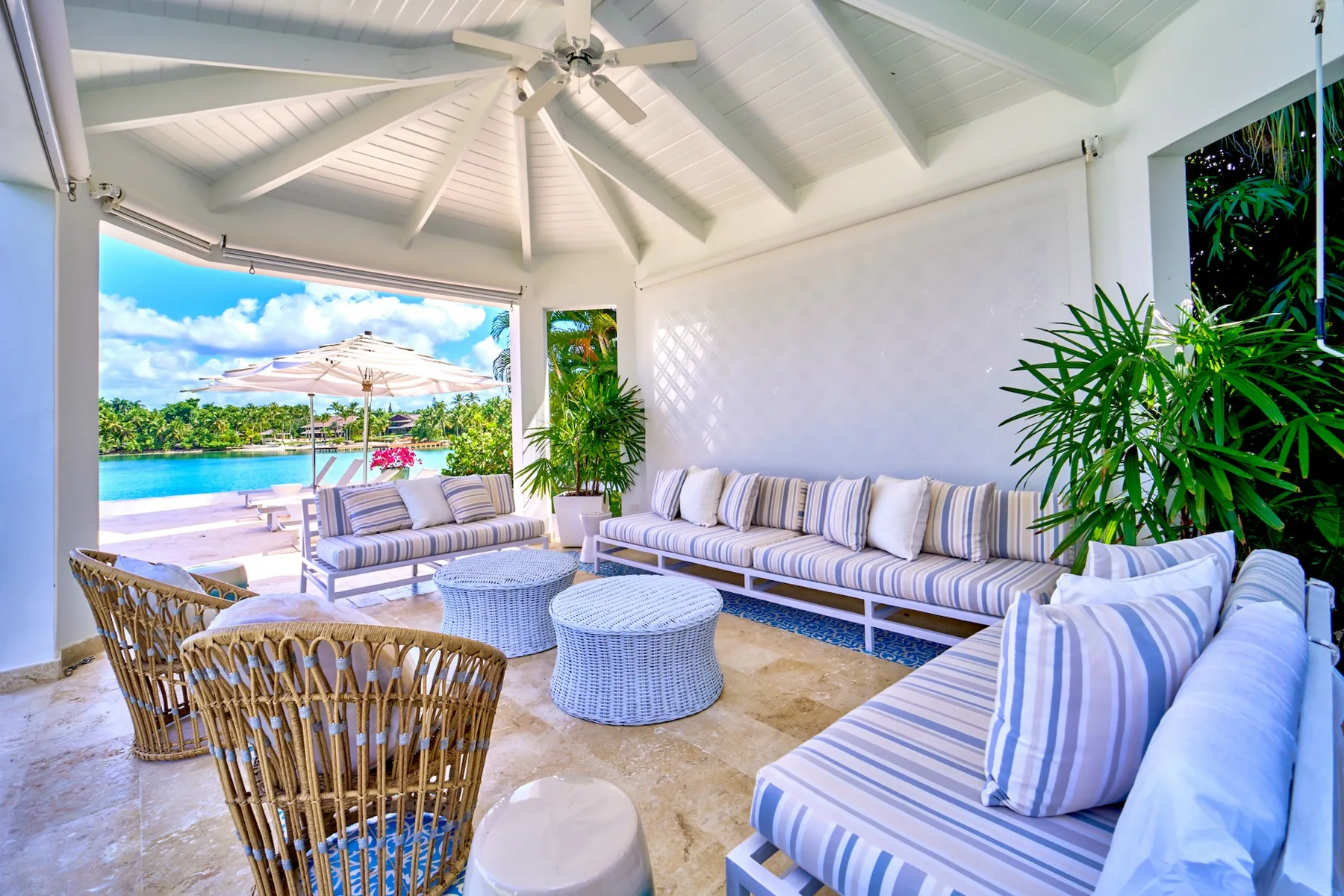 A bright covered lounge opens onto a poolside patio, with striped seating and leafy plants.