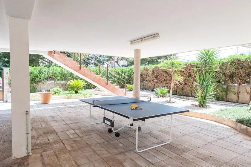 A covered patio has a ping-pong table, tiled flooring, and garden plants.
