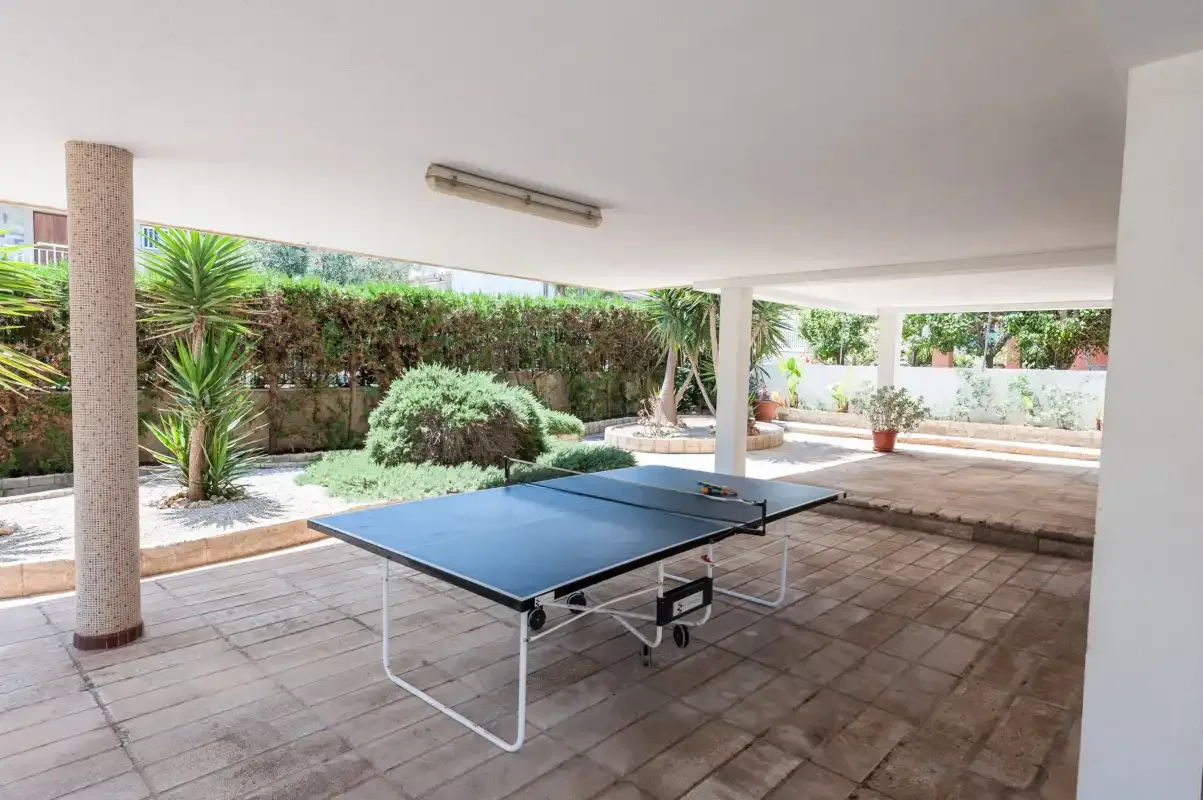 A covered outdoor patio has a table tennis table beside a planted garden.