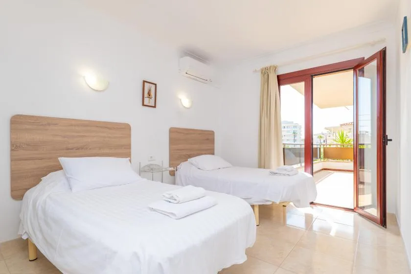 A bright bedroom with two beds and an open glass door to a balcony.
