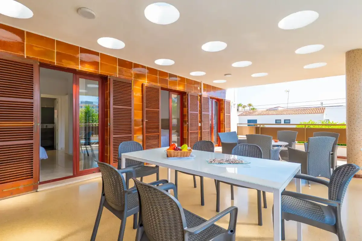 An outdoor dining area has a long table and woven seating beside shuttered windows.