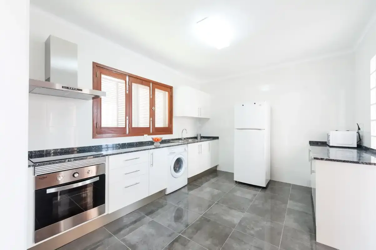 A bright kitchen with white cabinets, a refrigerator, and gray tiled flooring.