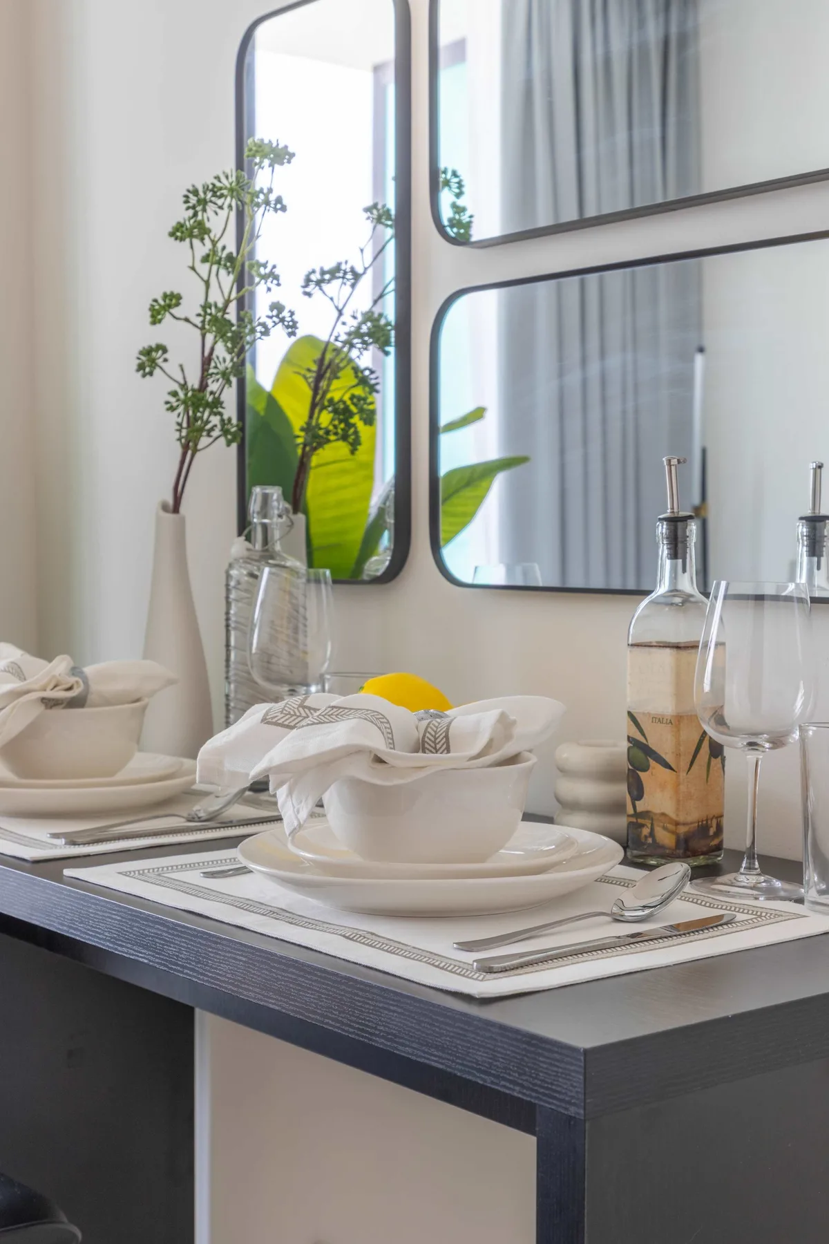 A dining table is set with white dishes, glassware, and a small vase beside a window.