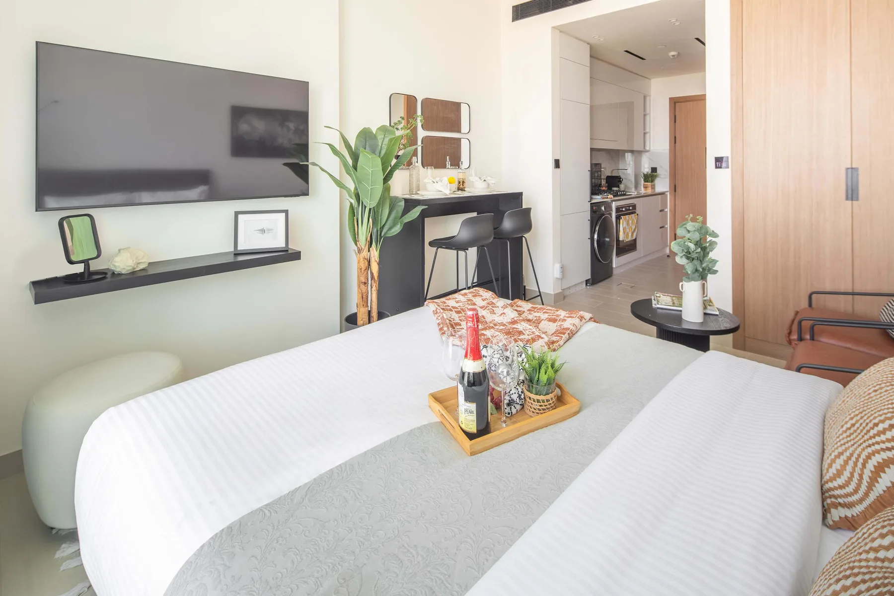 A bright bedroom with a bed, wall-mounted television, and kitchenette in the background.