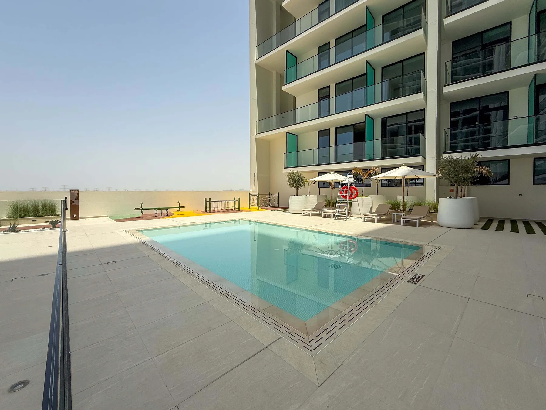 An outdoor swimming pool sits beside a modern building with balconies and lounge seating.