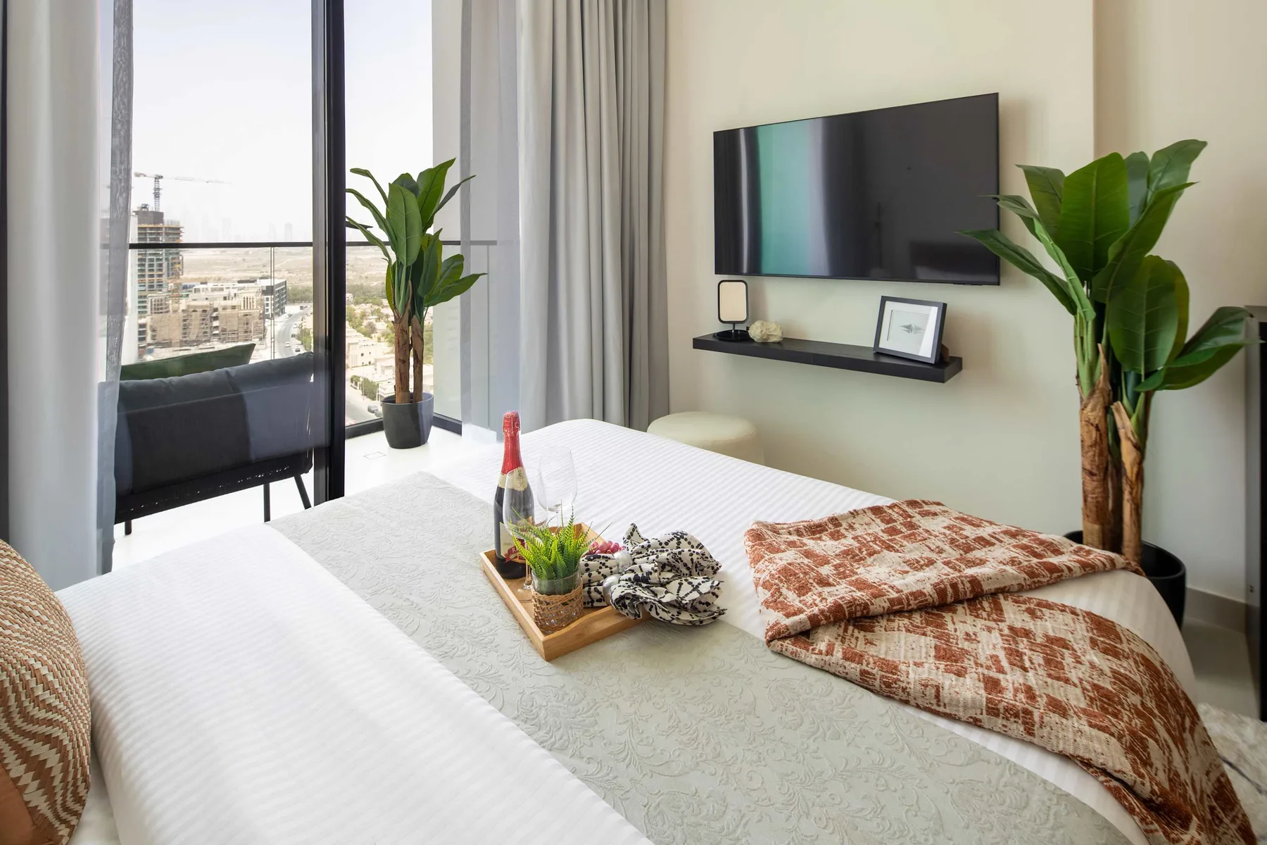 A bedroom with a neatly made bed, wall-mounted television, and plants beside a glass door.