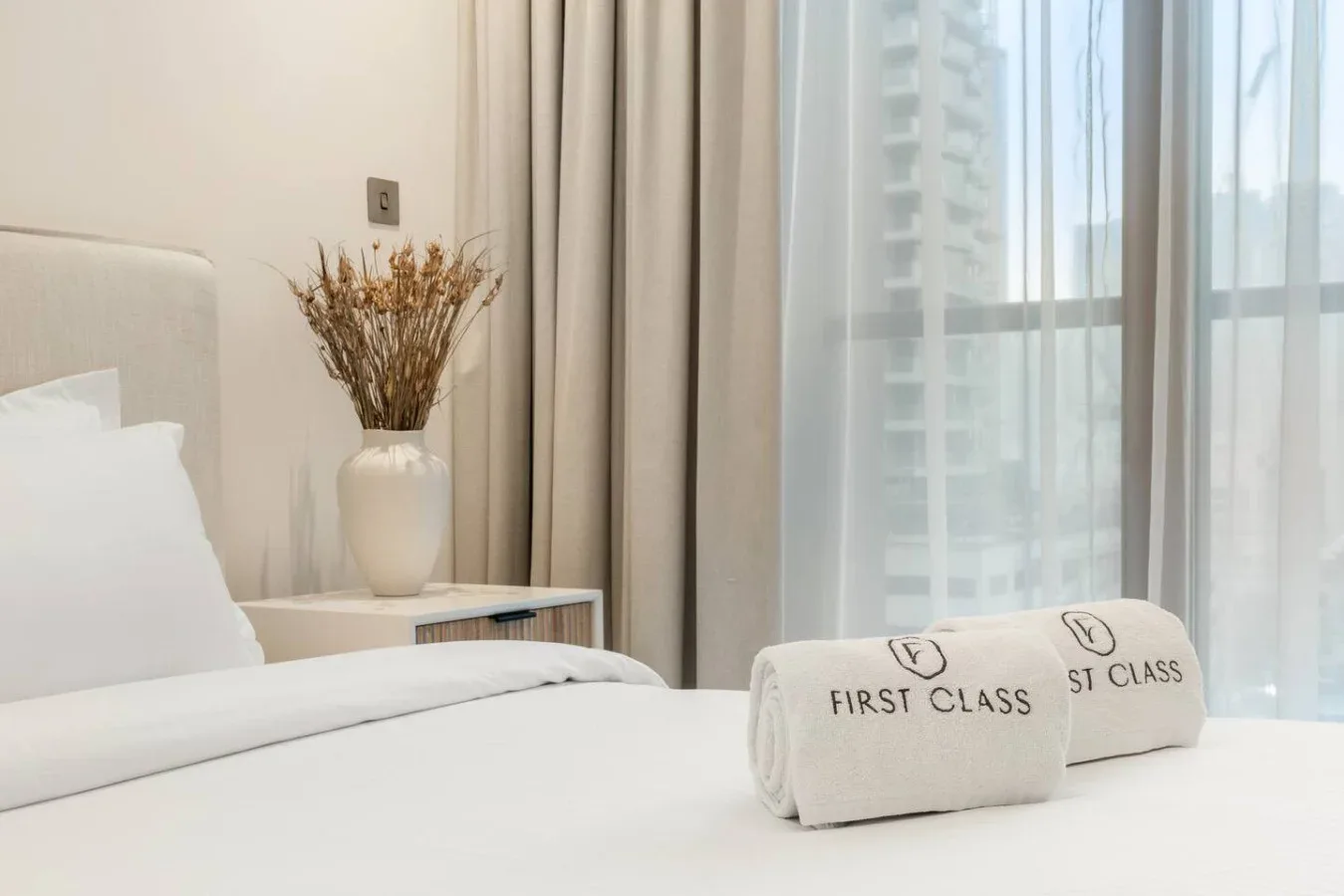 Un dormitorio ordenado con ropa de cama blanca, flores junto a la cama y cortinas ante una ventana luminosa.
