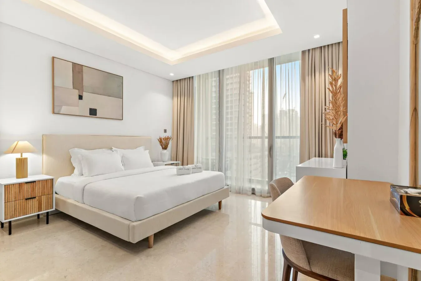 Un dormitorio luminoso con una cama bien tendida y una mesa junto a ventanas altas.