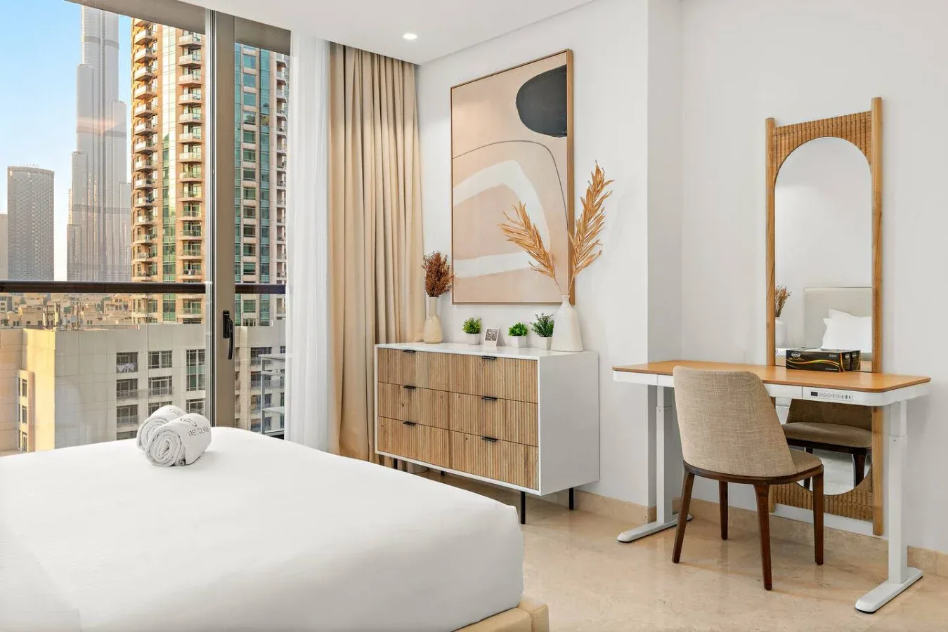 Un dormitorio luminoso tiene una cama, una cómoda de madera y un escritorio junto a grandes ventanas.