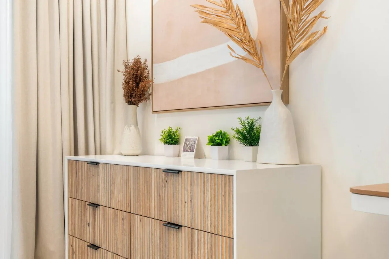 Un mueble de madera clara está bajo un cuadro abstracto, con plantas pequeñas y jarrones encima.