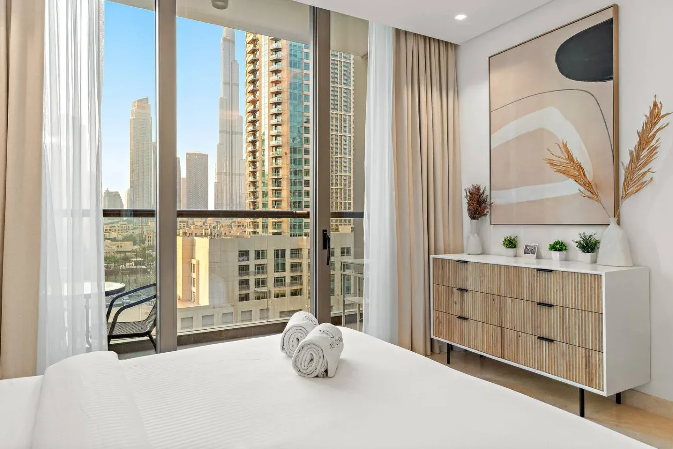 Un dormitorio luminoso con una cama bien arreglada junto a grandes ventanales con vistas a edificios.