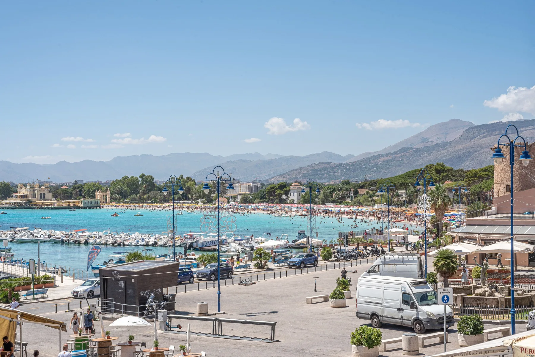 Un concurrido paseo marítimo da a una playa llena de gente y una bahía turquesa.