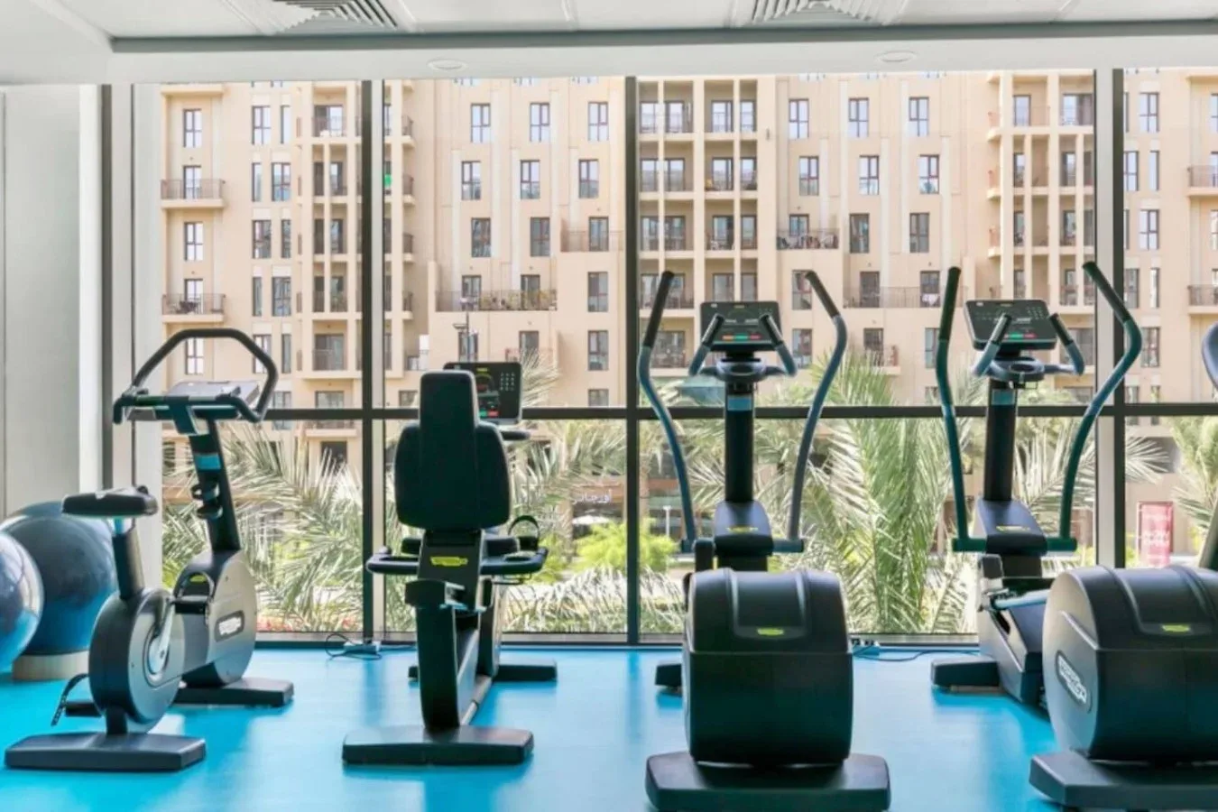 A fitness room with exercise bikes, treadmills, and large windows overlooking nearby buildings.