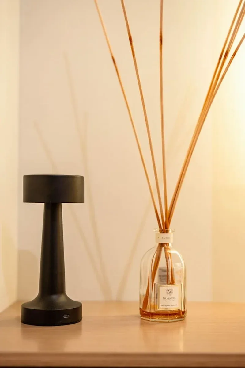 A table lamp and reed diffuser sit on a wooden tabletop against a plain wall.