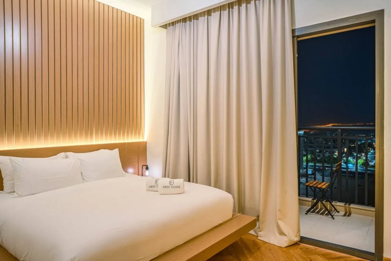 A bedroom with a neatly made bed, curtains, and a balcony overlooking city lights.
