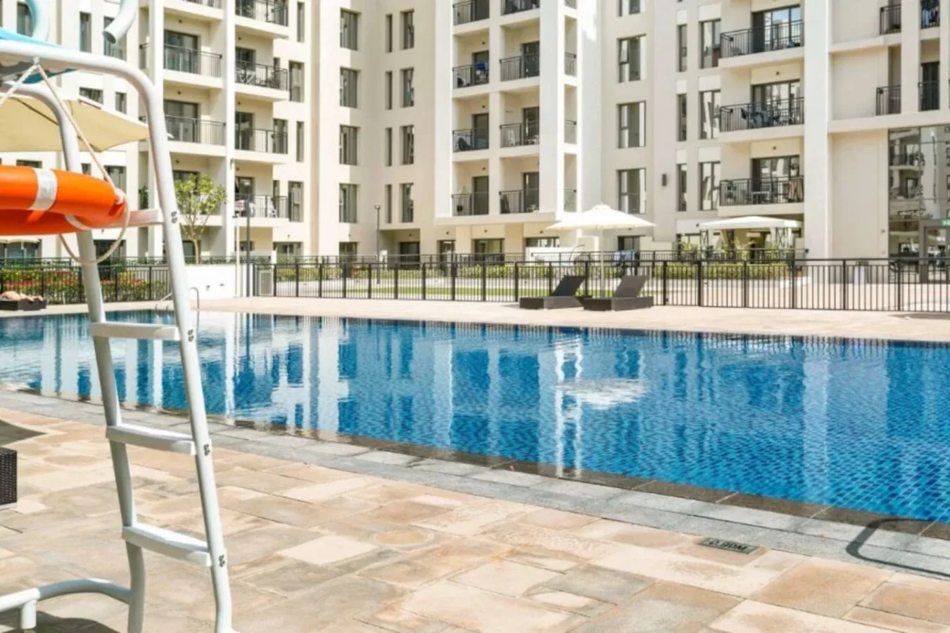 An outdoor swimming pool sits beside a paved deck and apartment buildings.