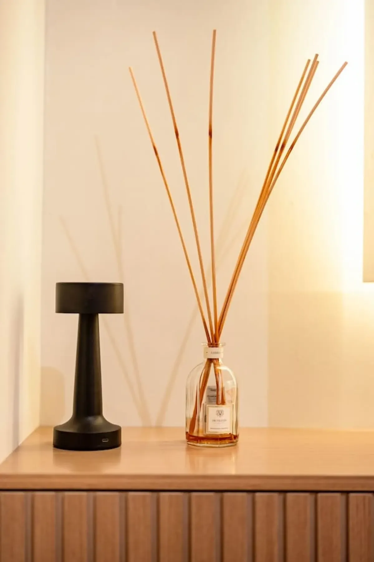 A black table lamp and reed diffuser sit on a wooden console.