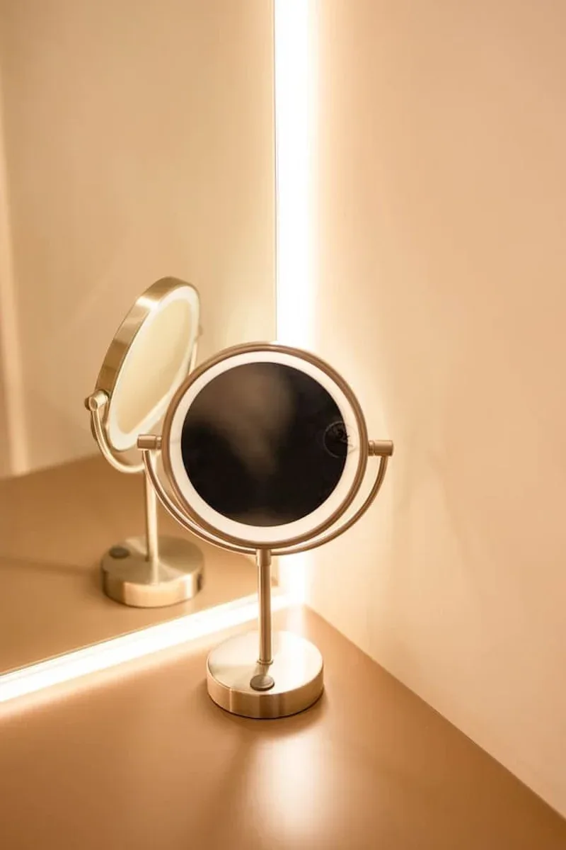 A round tabletop mirror sits on a counter beside a lit vertical strip.