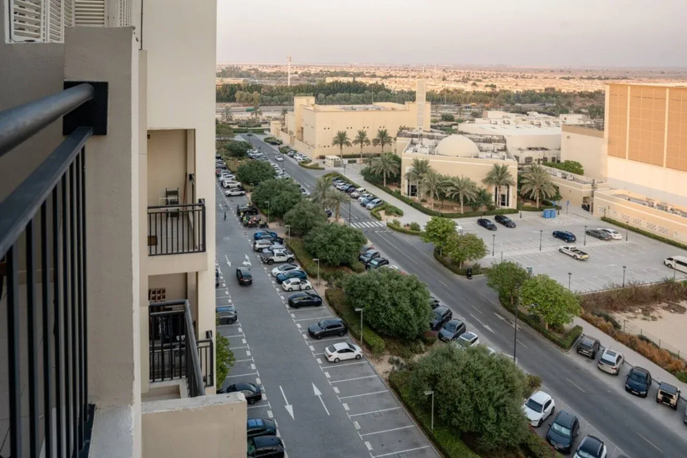 A balcony overlooks a tree-lined road, parking areas, and low-rise buildings.