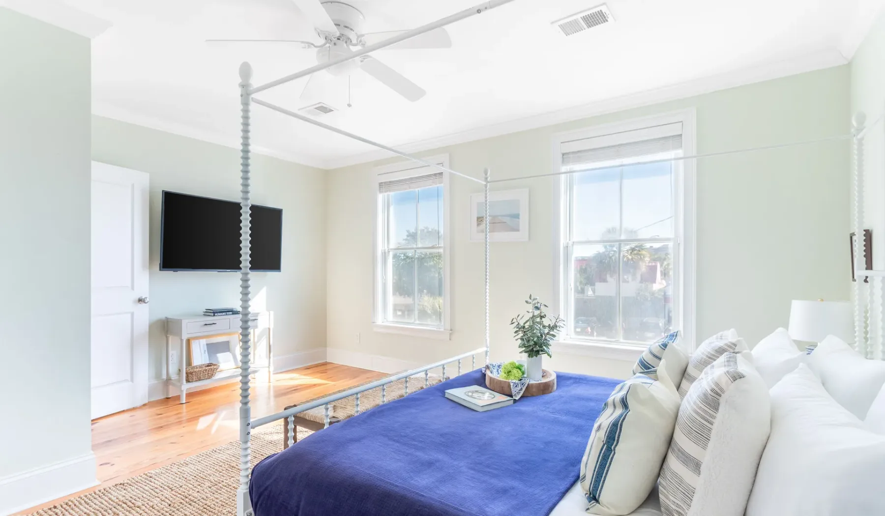 A bright bedroom with a blue bedspread, white seating, and sunlit windows.
