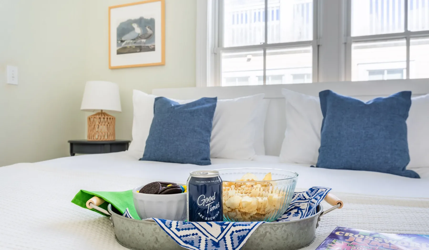 A bright bedroom features a neatly made bed with blue pillows and a tray of snacks.