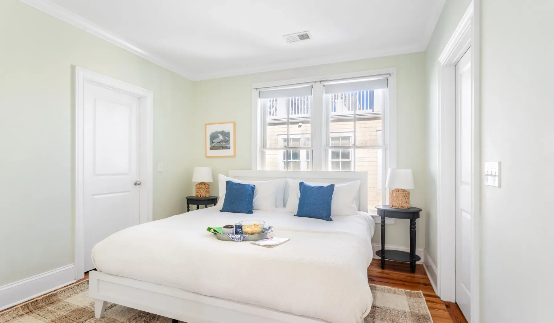 A bright bedroom with a white bed and blue pillows beneath two windows.