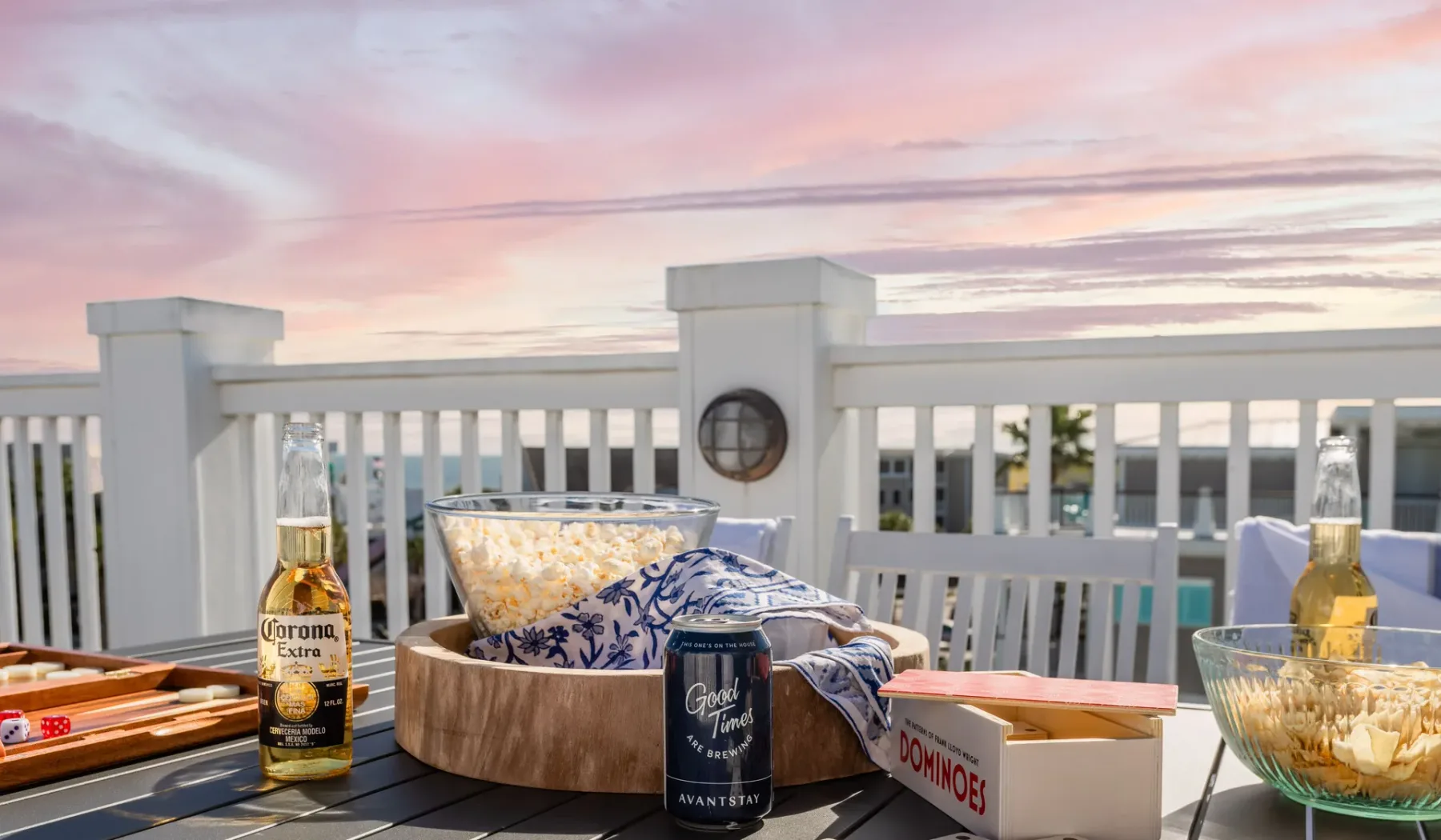 A deck table with snacks and drinks sits beside a white railing at sunset.