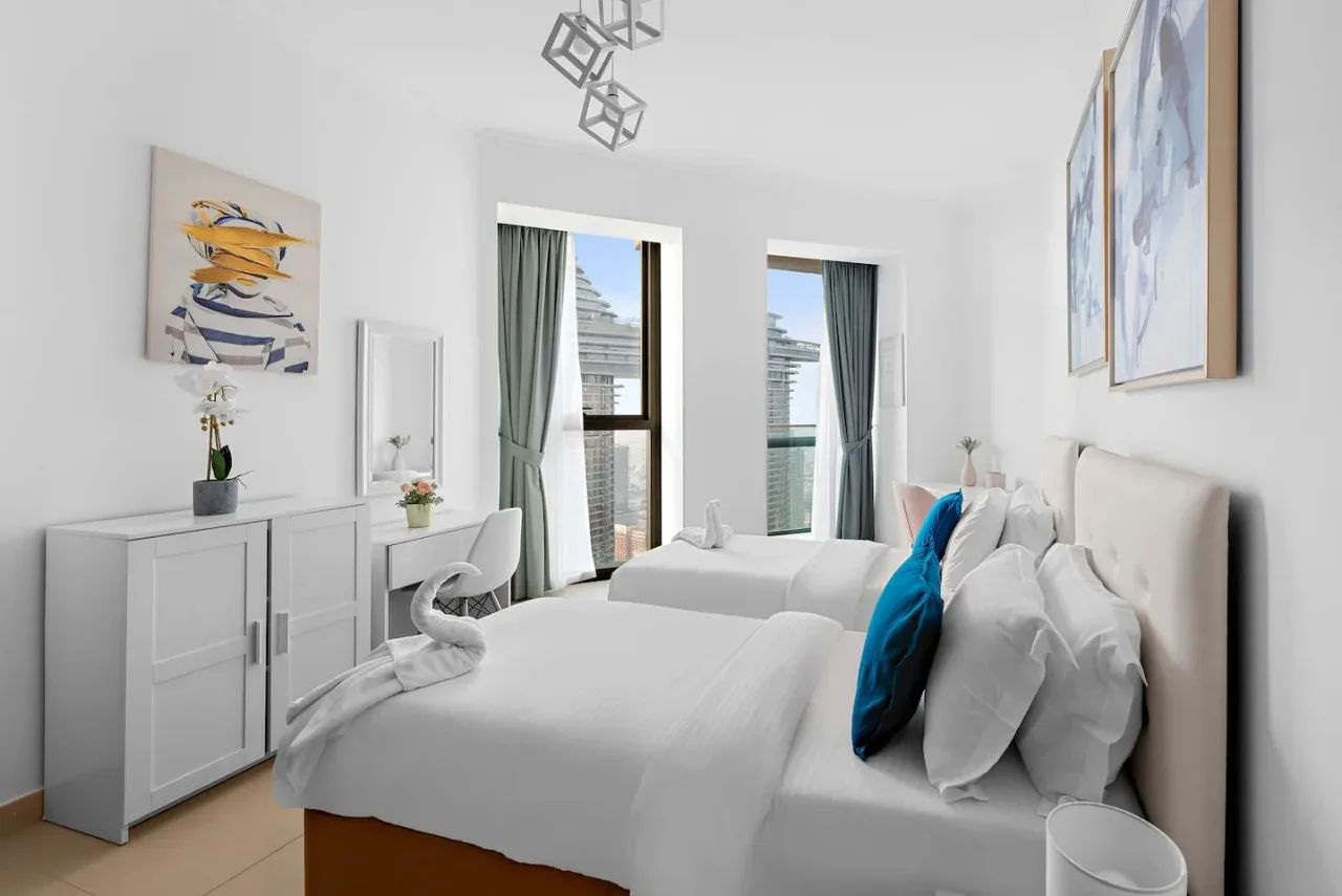 Un dormitorio luminoso con una cama blanca y grandes ventanas con vista a edificios cercanos.