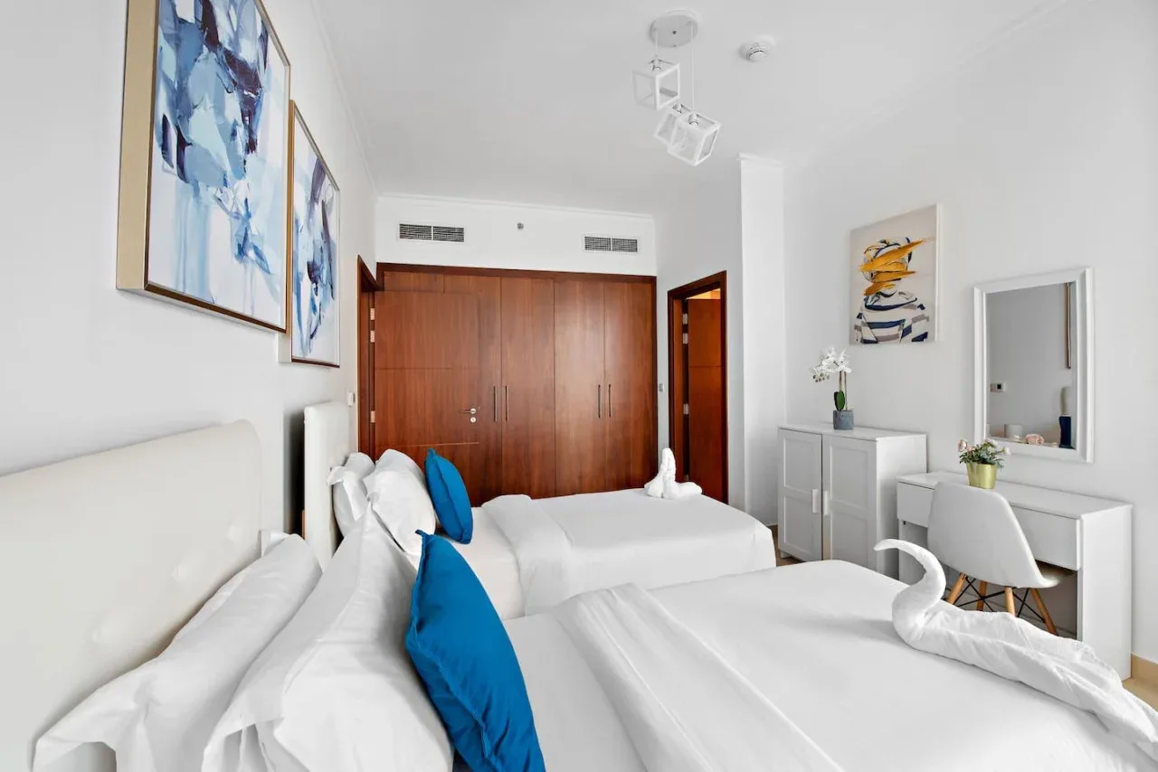 Un dormitorio luminoso con ropa de cama blanca, cojines azules y un armario de madera.