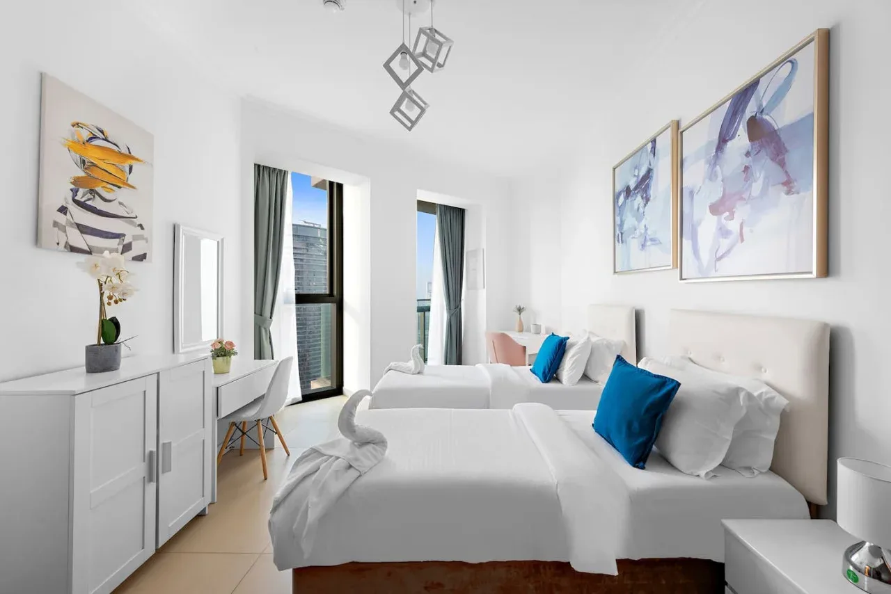Un dormitorio luminoso con dos camas, ropa de cama blanca y cuadros coloridos.