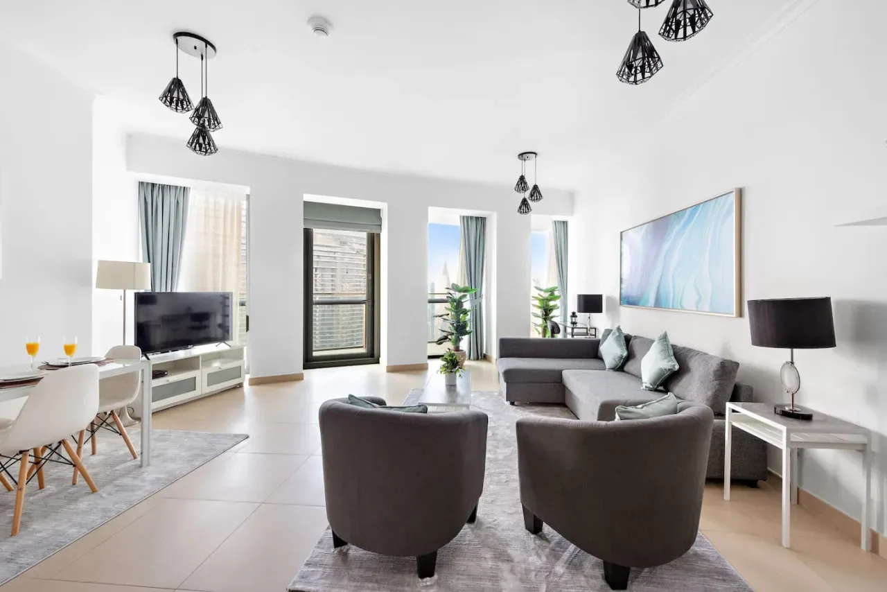 Una sala luminosa con asientos grises, un televisor y ventanas grandes.
