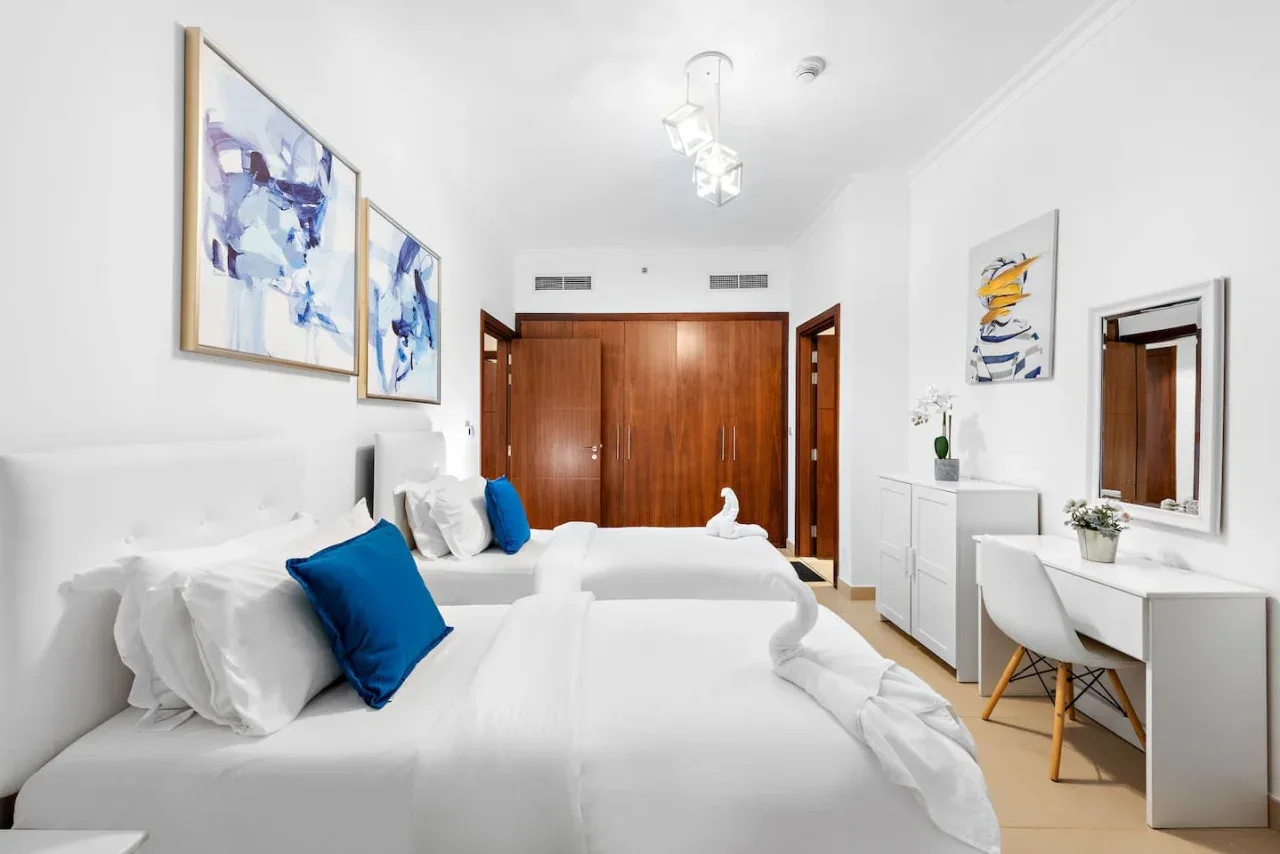 Un dormitorio luminoso con camas blancas, arte abstracto y una pequeña zona de tocador.