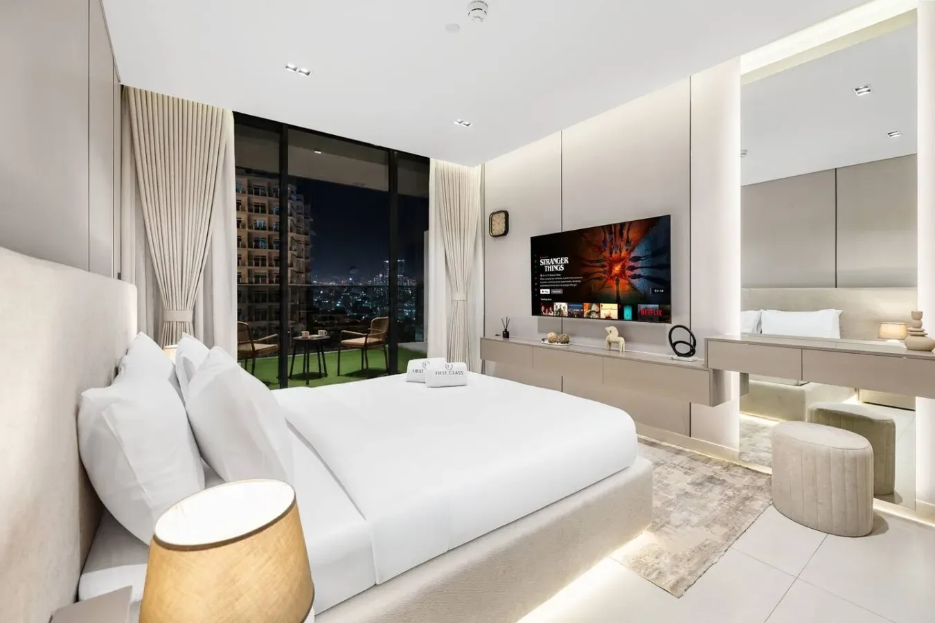 Un dormitorio luminoso con una cama blanca, un televisor y grandes ventanales.