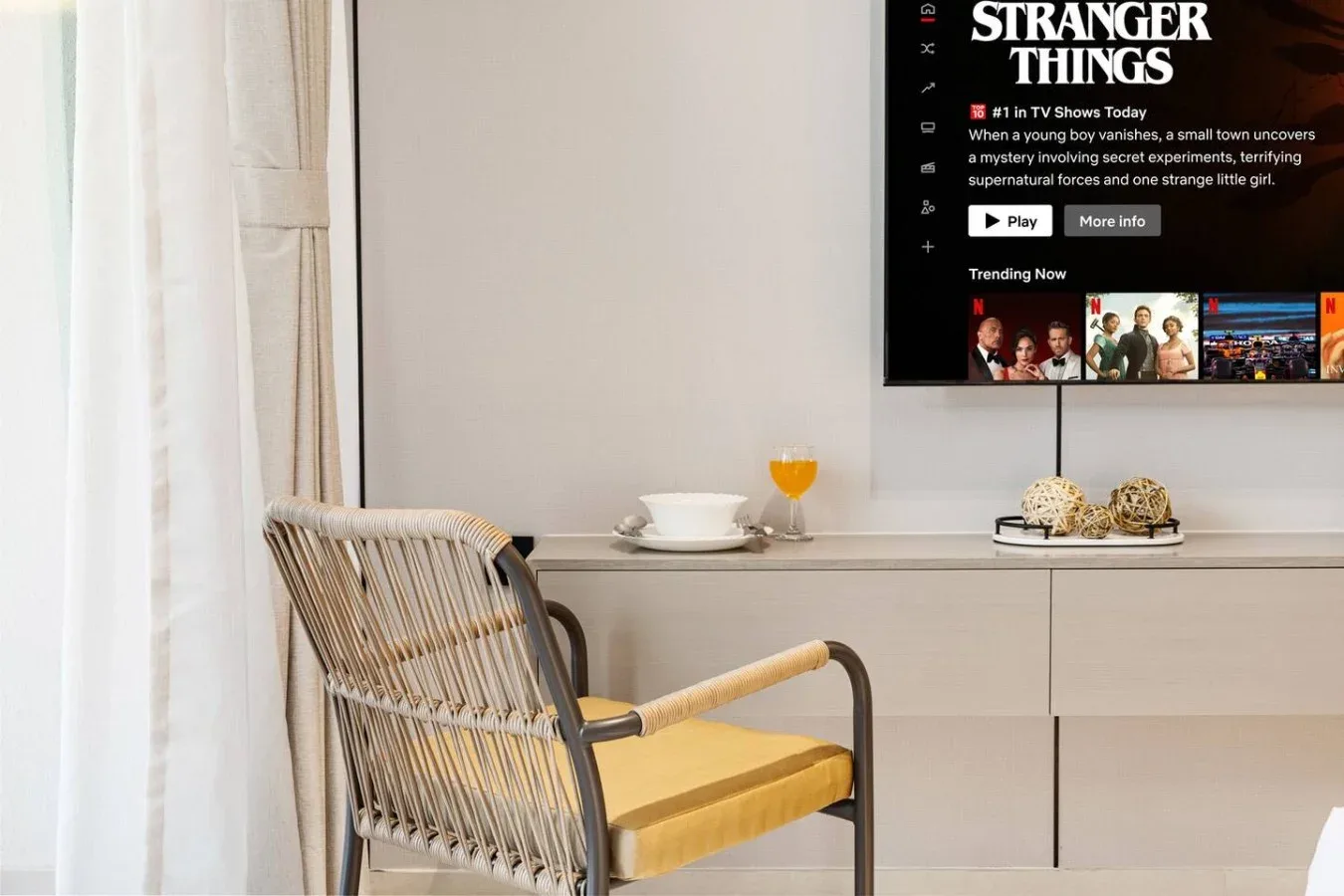 Una habitación de tonos claros con una silla tejida junto a un televisor montado en la pared.