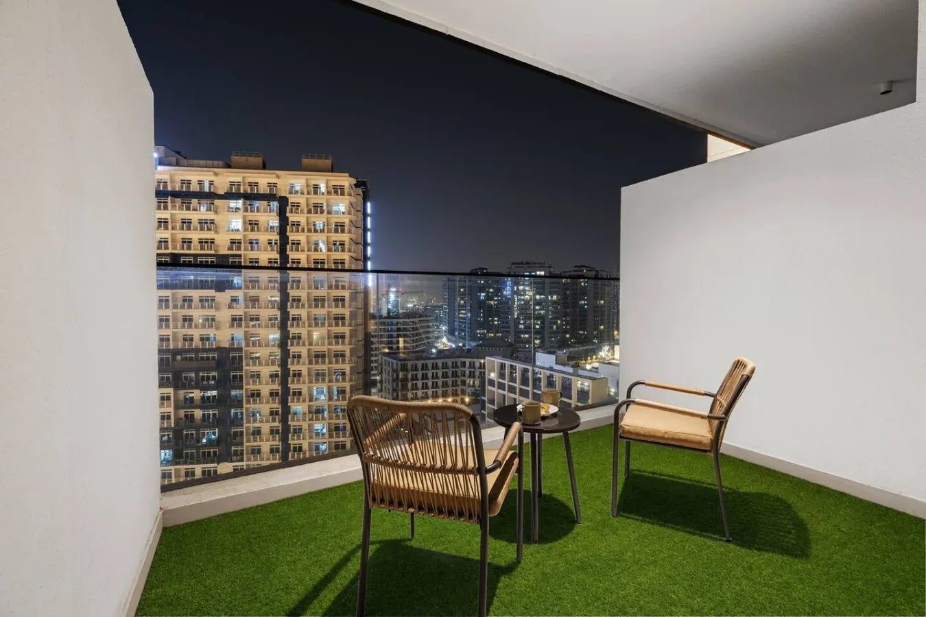 Un balcón con asientos y césped artificial tiene vistas a edificios iluminados de noche.