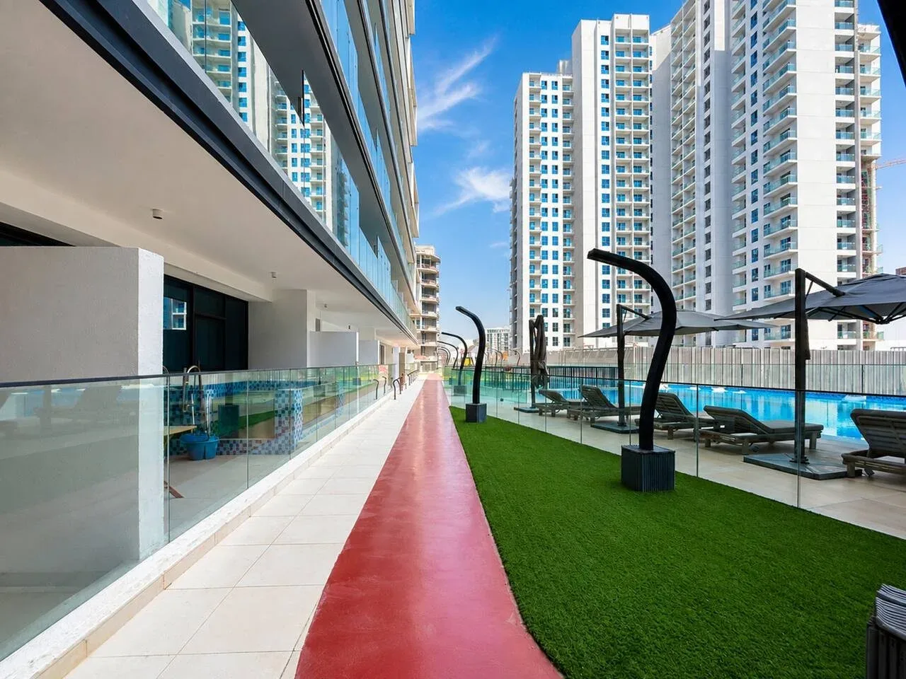 Un camino rojo bordea un césped verde y una piscina entre edificios altos.