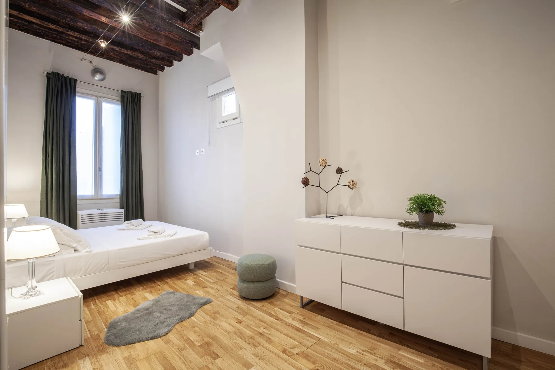 A bright bedroom with a bed, white dresser, and dark exposed ceiling beams.