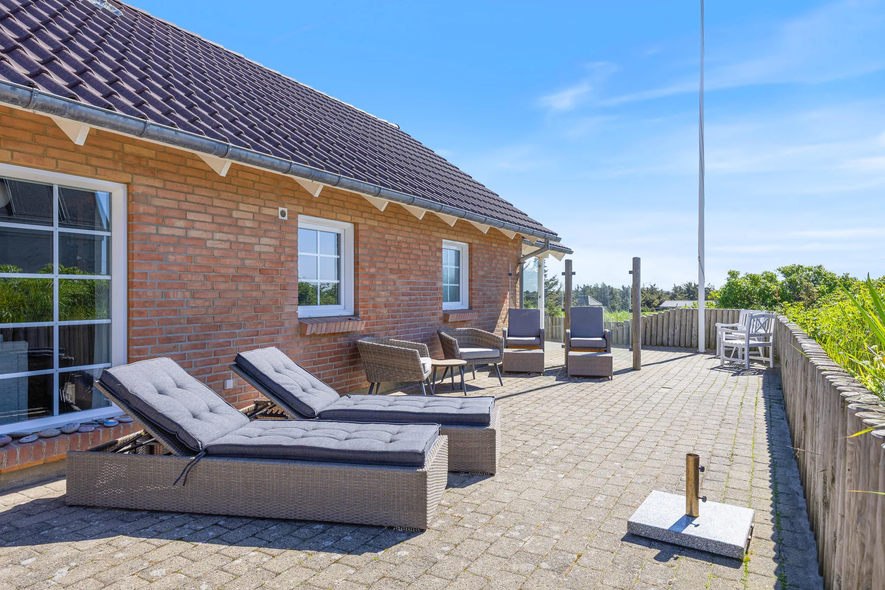 Un patio soleado y pavimentado junto a una casa de ladrillo tiene asientos al aire libre y una barandilla de madera.