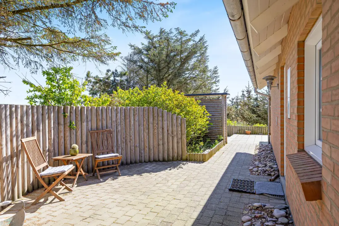 Un patio pavimentado junto a una pared exterior de ladrillo, con asientos de madera y un jardín cercado.