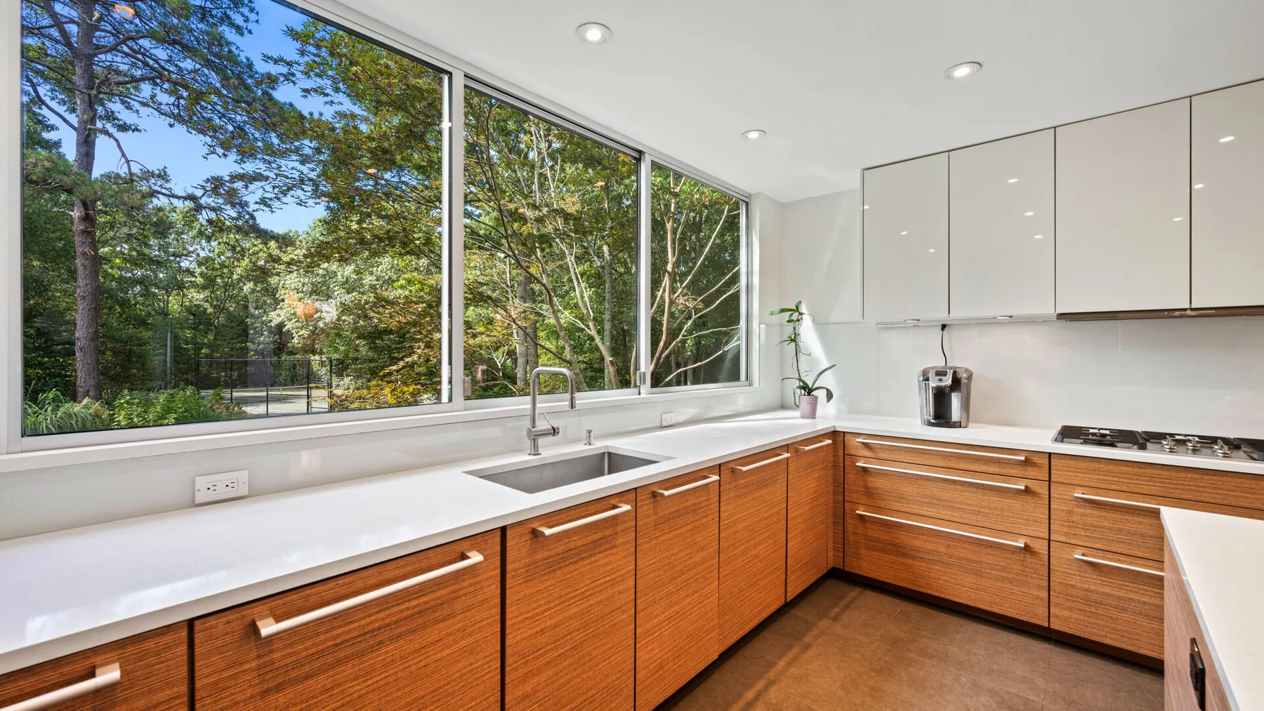 Una cocina luminosa tiene gabinetes de madera y amplias ventanas con vista a los árboles.