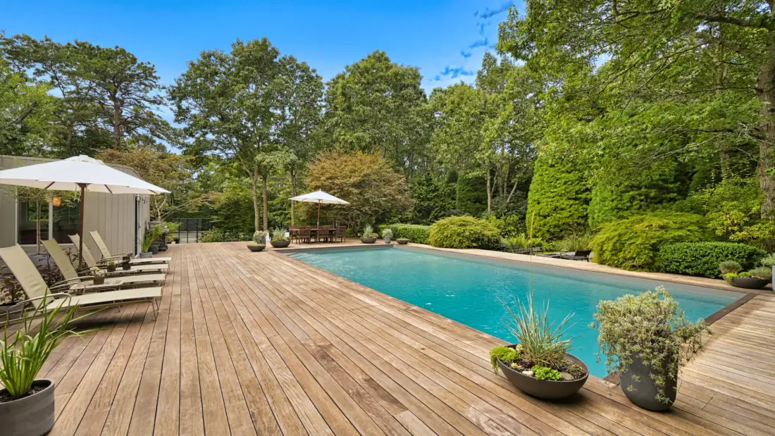Una piscina al aire libre junto a una amplia terraza de madera y árboles.
