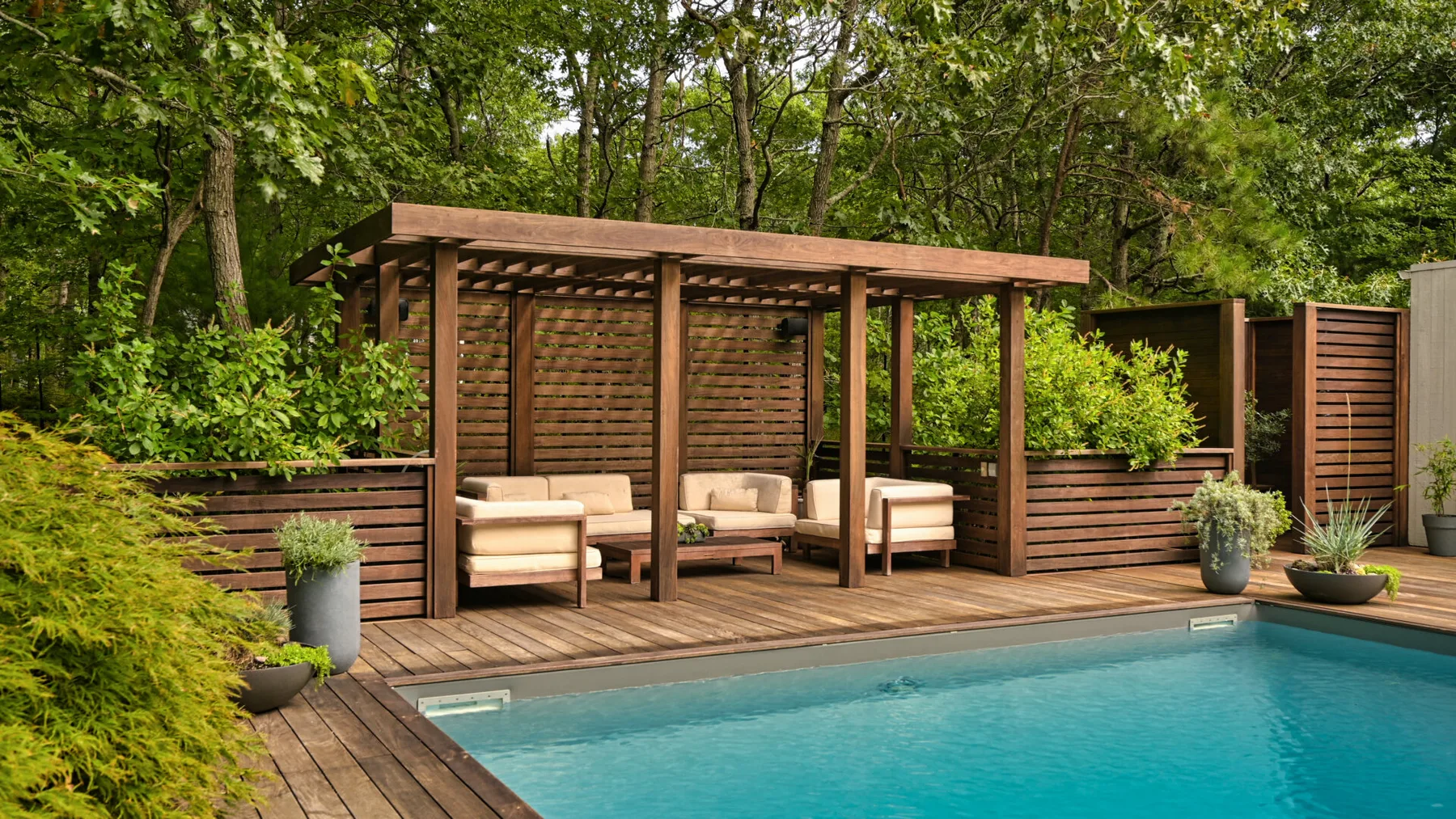 Una piscina al aire libre con pérgola de madera y asientos, rodeada de vegetación.