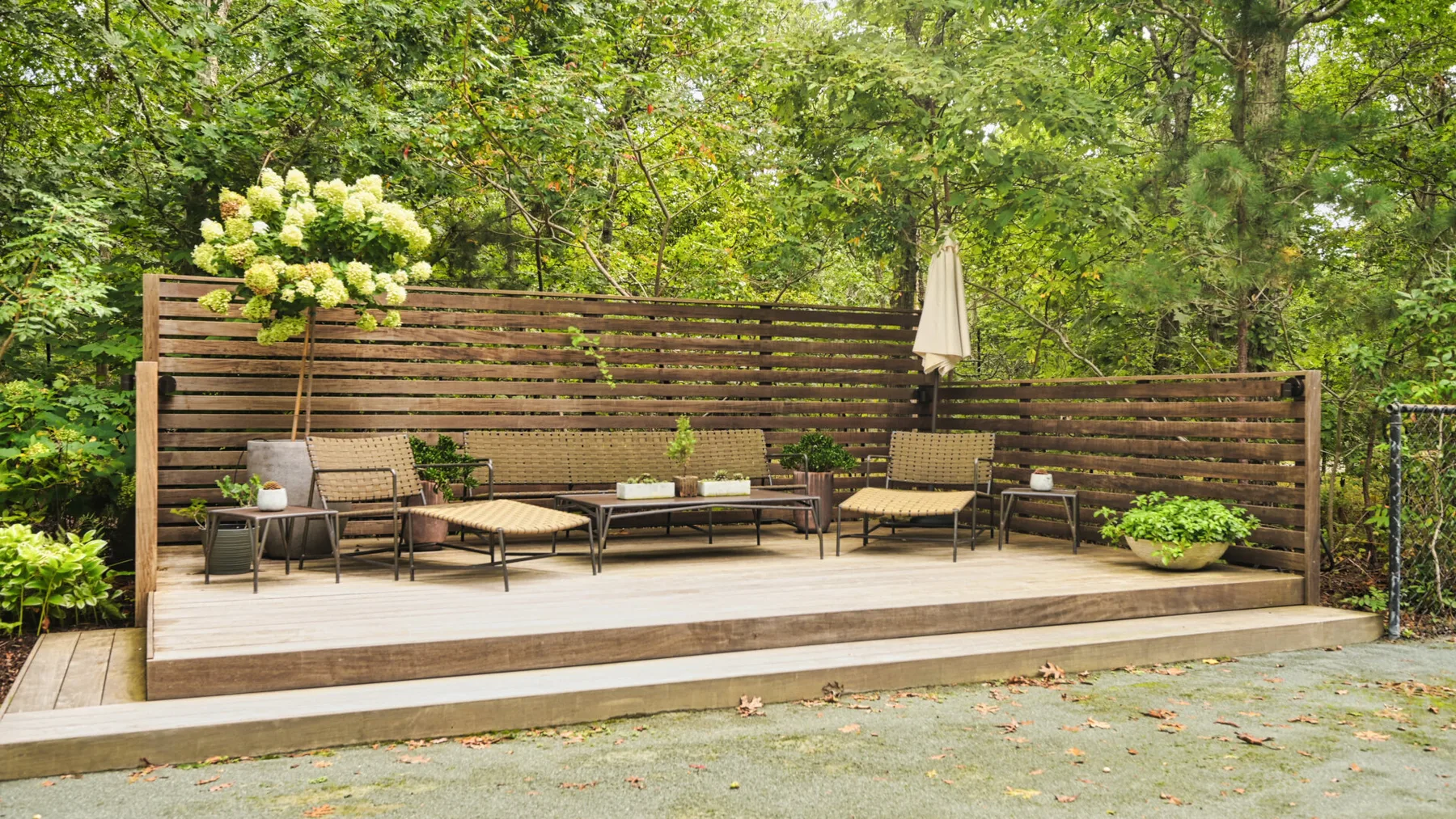 Una terraza exterior de madera tiene asientos, una mesa y vegetación alrededor.