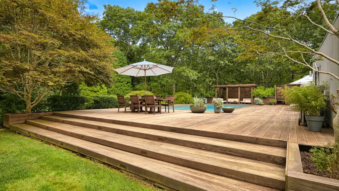 Una terraza de madera con escalones amplios, asientos y una sombrilla está rodeada de árboles.