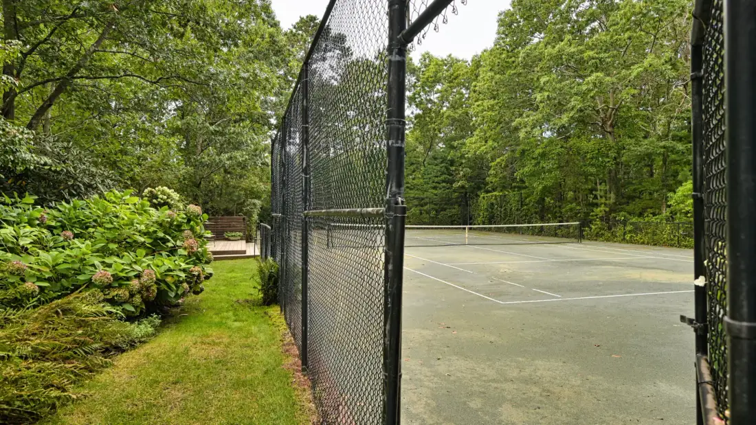 An outdoor tennis court is enclosed by a tall fence and surrounded by trees.