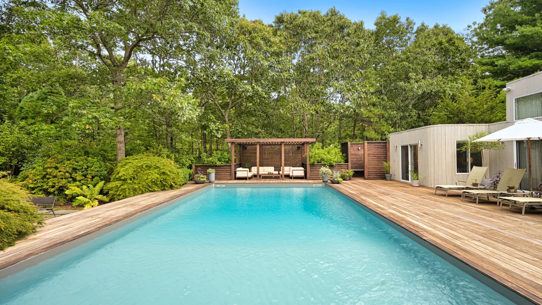 Una piscina al aire libre está junto a una terraza de madera y árboles frondosos.