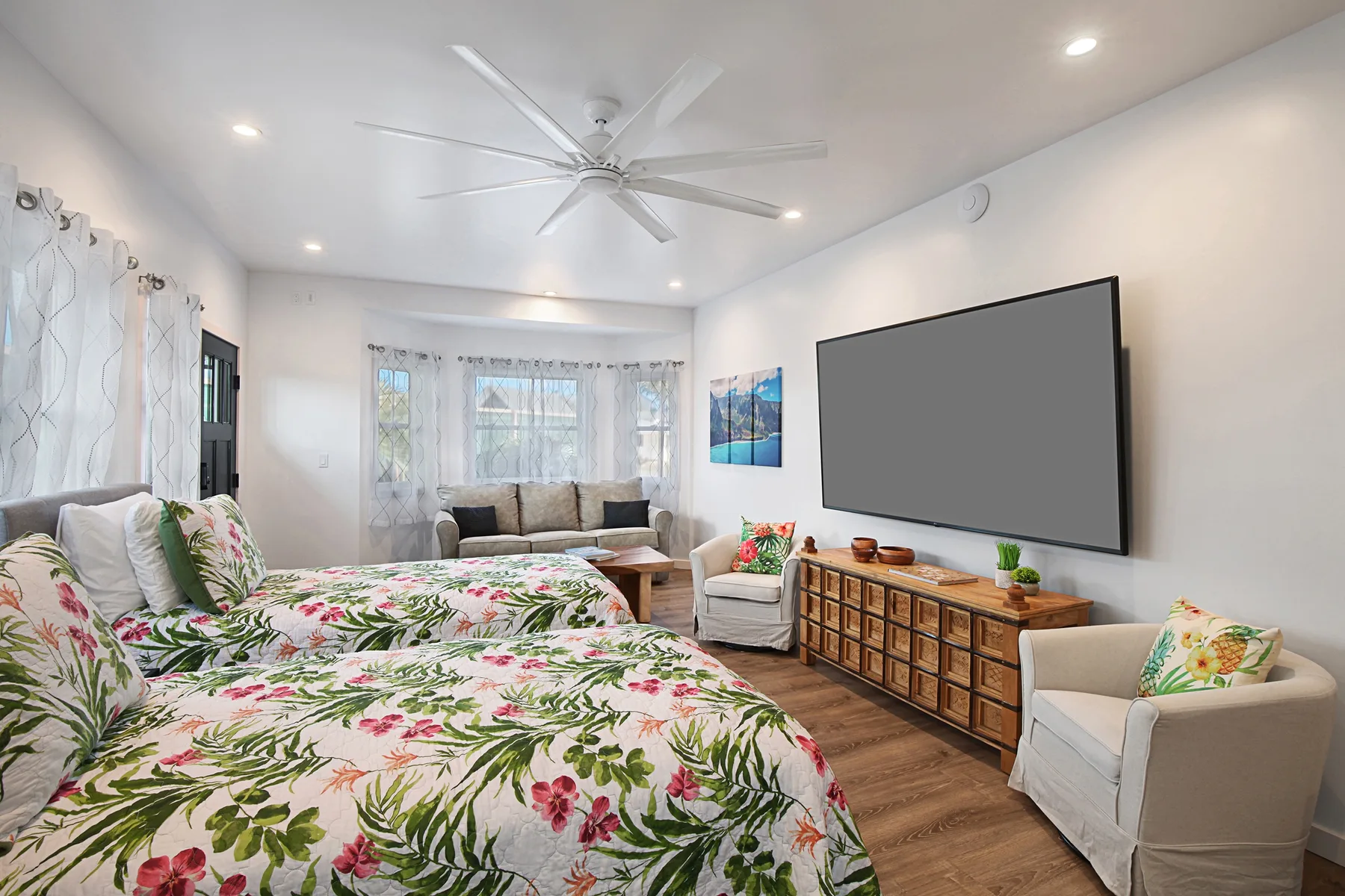 A bright bedroom with floral bedding, seating, and a large wall-mounted television.