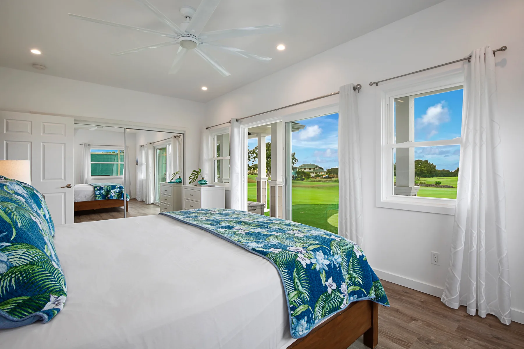 A bright bedroom with a large bed, blue patterned bedding, and windows overlooking a green lawn.