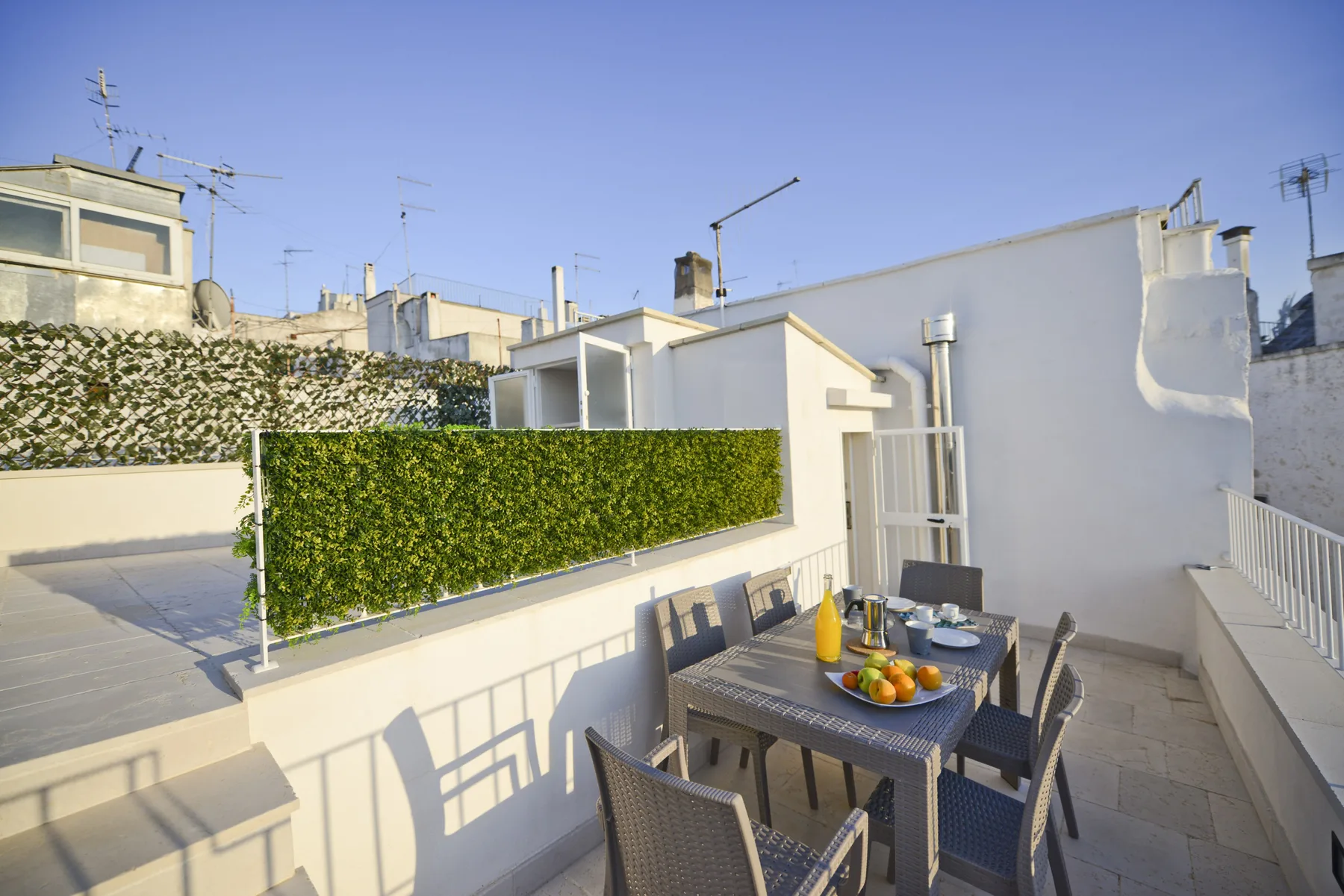 A rooftop dining terrace with a table and chairs beside a green hedge.